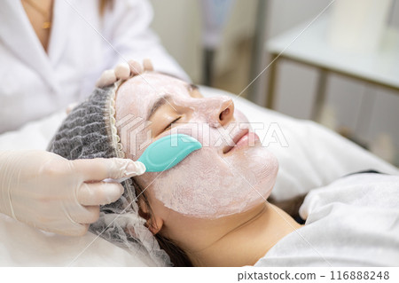 Cosmetologist applying a clay face mask with a blue spatula on a smiling young woman's face. Cosmetologist applying a clay face mask with a blue spatula on a smiling young woman's face. 116888248