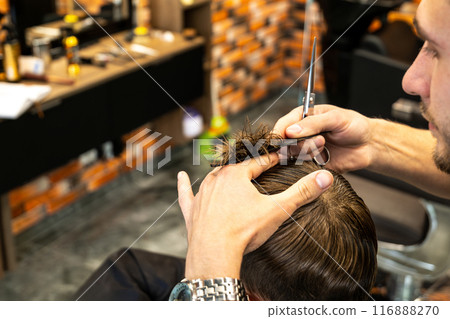 Young male barber making a haircut for cute little boy in the modern barbershop. Child in barbershop. Haircuts for Kids 116888270