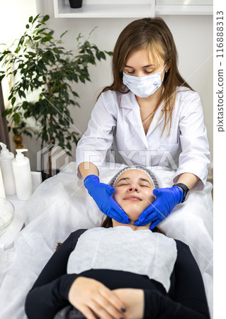 Beautician making face massage, applying cream or gel, with her hands in rubber gloves, to client's face in beauty clinic. The client is lying down o a treatment table relaxing and smiling. Beautician making face massage, applying cream or gel, with her hands in rubber gloves, to client's face in beauty clinic. The client is lying down o a treatment table relaxing and smiling. 116888313