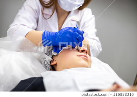 Close-up shot of a beautiful young woman on a facial derma pen micro-needling treatment at the beauty salon. The cosmetologist is wearing a face mask and medical gloves. Close-up shot of a beautiful young woman on a facial derma pen micro-needling treatment at the beauty salon. The cosmetologist is wearing a face mask and medical gloves. 116888316