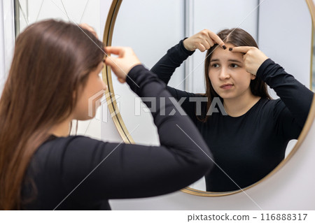 Young, beautiful woman in the dermatologist's cabinet looks at herself in the mirror, feeling disappointed with the quality of her skin. Young, beautiful woman in the dermatologist's cabinet looks at herself in the mirror, feeling disappointed with the quality of her skin. 116888317