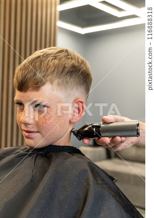 Head cut in the barbershop. The barber cuts the hair from the client's head. The process of creating men's hairstyles. Hairdresser's. Blond boy with freckles on his face and alabaster eyes. Head cut in the barbershop. The barber cuts the hair from the client's head. The process of creating men's hairstyles. Hairdresser's. Blond boy with freckles on his face and alabaster eyes. 116888318