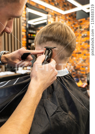 The picture of a cute little child with blonde hair in the process of being cut. A professional in the field makes a line at the child's head. The picture of a cute little child with blonde hair in the process of being cut. A professional in the field makes a line at the child's head. 116888319