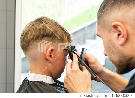 Close-up of man hands grooming kid boy hair in barber shop. Boy cut with hairdresser's machine. Portrait of male child at the barber shop to cut his hair. 116888329