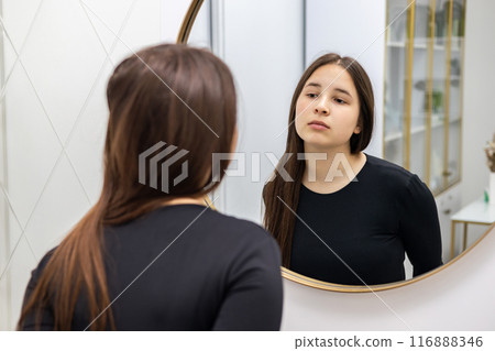 Beautiful young woman with long hair, wearing a black blouse, looking in the mirror unsatisfied, getting ready for a skin treatment. 116888346