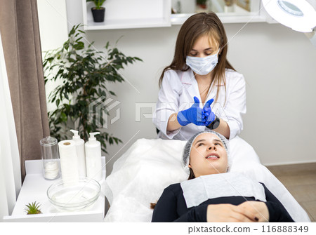 Close-up of beautician using her hands in sterile gloves while applying facial jelly gel to female client's face in beauty clinic. Applying a solution to the face, cosmetic procedure. Close-up of beautician using her hands in sterile gloves while applying facial jelly gel to female client's face in beauty clinic. Applying a solution to the face, cosmetic procedure. 116888349