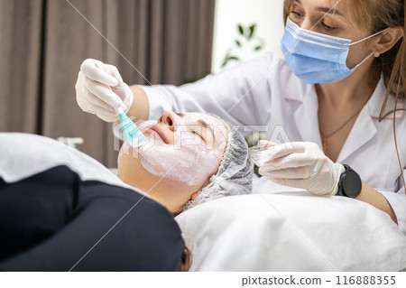 Cosmetologist wearing a surgical mask and gloves, applying a face mask with a spatula to her client Cosmetologist wearing a surgical mask and gloves, applying a face mask with a spatula to her client 116888355