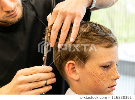 Children hairdresser with scissors is cutting little boy against a dark background. Contented cute preschooler boy getting the haircut.  116888391