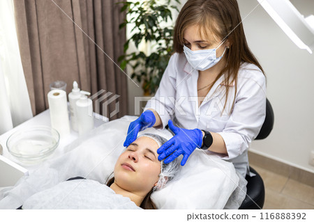 Facial massage is prepared in the beauty salon. Doctor's hands. Treatment for healthy skin. Head of a young woman. Cleansing rejuvenation. Dermatological face mask. Detox therapy 116888392