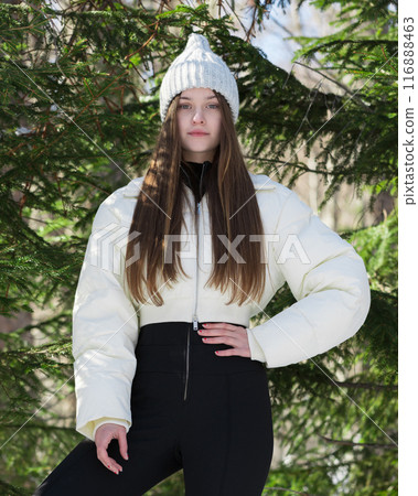 Gorgeous teenage girl stands in winter pine woodland and gazing at camera. Female fashion model is dressed to nines in cozy outdoor attire, including knitted hat, white puffer down jacket, black pants Gorgeous teenage girl stands in winter pine woodland and gazing at camera. Female fashion model is dressed to nines in cozy outdoor attire, including knitted hat, white puffer down jacket, black pants 116888463