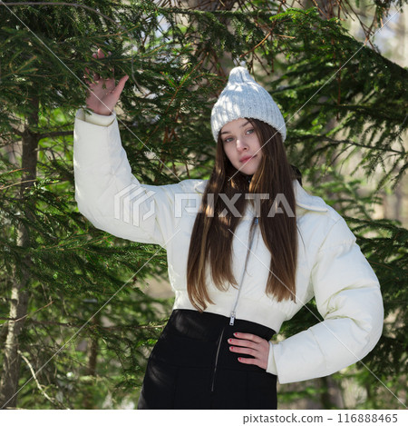 Teenage girl stands proudly in winter pine forest. She's brunette, looking absolutely radiant as she gazes into camera. Dressed to impress, fashion model in knitted hat, white puffer bomber jacket Teenage girl stands proudly in winter pine forest. She's brunette, looking absolutely radiant as she gazes into camera. Dressed to impress, fashion model in knitted hat, white puffer bomber jacket 116888465