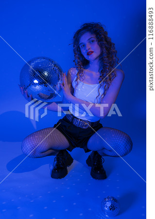 Young woman exuding sensuality is striking poses in squatting position while having blast at nightclub with mesmerizing disco ball. Blue neon colored lights add an extra touch of excitement to scene 116888493