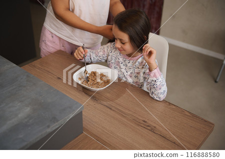A young girl eating cereal for breakfast while a person helps her. She is seated at a wooden table, enjoying her morning meal. 116888580