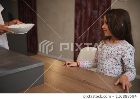 A young girl wearing floral pajamas eagerly waiting at the kitchen table while someone serves her food. Capturing the anticipation and excitement of mealtime. 116888594