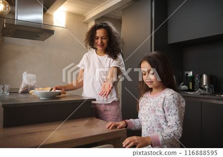 A mother and her daughter spend quality time together in a cozy kitchen. They are preparing breakfast and enjoying their morning routine. A mother and her daughter spend quality time together in a cozy kitchen. They are preparing breakfast and enjoying their morning routine. 116888595