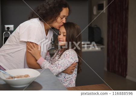 A tender moment between a mother and daughter as they share a hug and kiss in a cozy kitchen. Warm emotions of love, care, and family bond are depicted in this heartwarming scene. A tender moment between a mother and daughter as they share a hug and kiss in a cozy kitchen. Warm emotions of love, care, and family bond are depicted in this heartwarming scene. 116888596