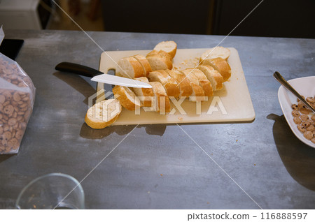 A close-up shot of freshly sliced French bread on a cutting board with a knife beside it. Nearby is a bowl with cereal on a kitchen counter. 116888597