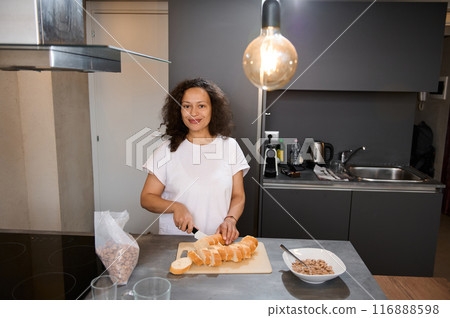 Smiling woman slicing fresh bread in a modern kitchen. She stands at a counter with a bowl of nuts and a cutting board, creating a homely and welcoming atmosphere. Smiling woman slicing fresh bread in a modern kitchen. She stands at a counter with a bowl of nuts and a cutting board, creating a homely and welcoming atmosphere. 116888598