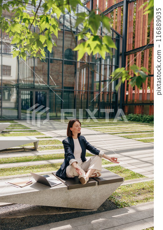 A woman is peacefully meditating outdoors on a bench in a modern office courtyard A woman is peacefully meditating outdoors on a bench in a modern office courtyard 116889035