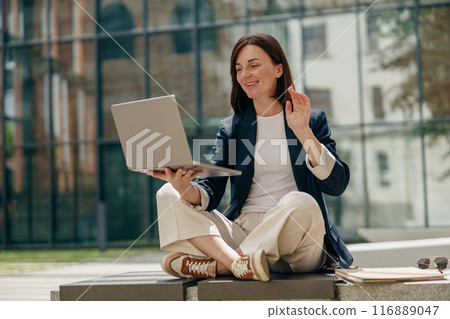 A businesswoman is outdoors waving during a video call with a laptop while a student is also present 116889047