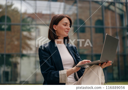 A businesswoman is focused on her laptop, working outside the corporate building 116889056