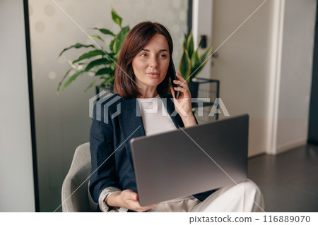 Confident businesswoman multitasking, working on laptop and talking on phone in a modern office 116889070
