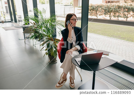 A businesswoman in modern office, focused and professional, works on laptop and phone 116889093