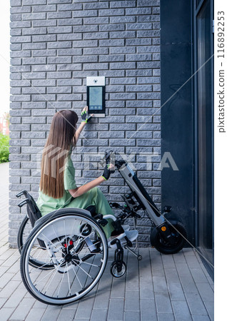 A woman in a wheelchair with an assistive device for manual control enters the entrance. Electric hand bike. A woman in a wheelchair with an assistive device for manual control enters the entrance. Electric hand bike. 116892253