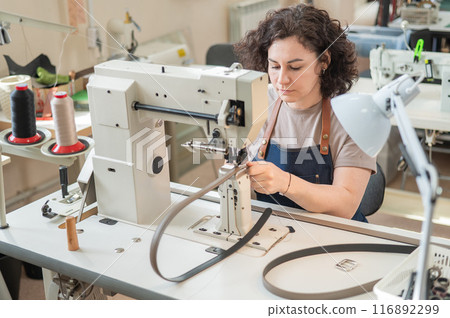 A woman tanner sews a leather belt on a sewing machine.  116892299