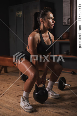 Caucasian woman doing squats with kettlebell in retro gym. Vertical photo. Caucasian woman doing squats with kettlebell in retro gym. Vertical photo. 116892313