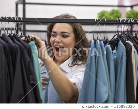 Portrait of a fat woman in a plus size store through hangers with clothes. Portrait of a fat woman in a plus size store through hangers with clothes. 116892320