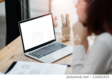 A woman working on a laptop with a blank screen at a wooden desk, surrounded by office supplies and documents, in a modern office environment. A woman working on a laptop with a blank screen at a wooden desk, surrounded by office supplies and documents, in a modern office environment. 116892425