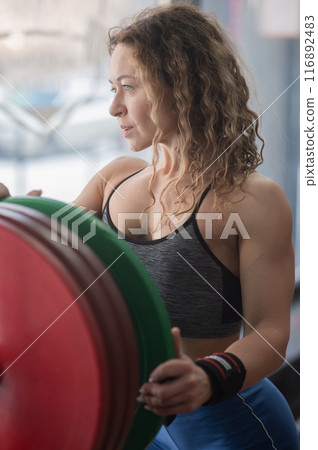 Caucasian forty year old woman putting a weight plate on a barbell in the gym. Vertical photo.  116892483