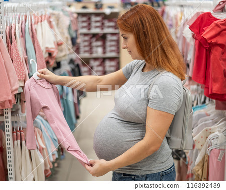 Caucasian pregnant woman chooses baby clothes in a store.  116892489