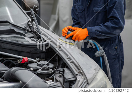 A mechanic sands the putty on a car body with a machine. Repair after an accident. A mechanic sands the putty on a car body with a machine. Repair after an accident. 116892742