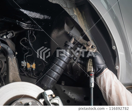 An auto mechanic applies anti-corrosion mastic to the underbody of a car. An auto mechanic applies anti-corrosion mastic to the underbody of a car. 116892782