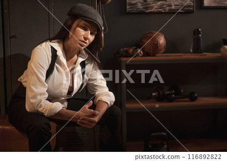 Caucasian woman in a Peaky Blinders style men's suit posing in a retro gym. 116892822