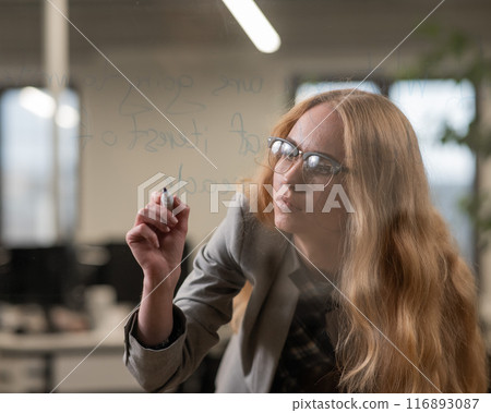 Caucasian woman with glasses writes text in English on a glass wall.  116893087