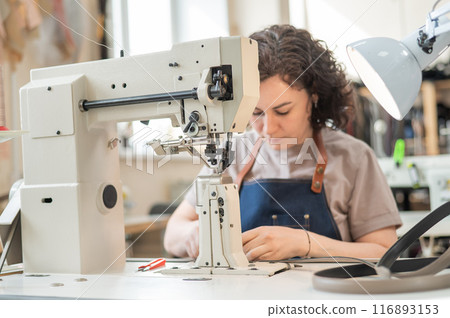 A woman tanner sews a leather belt on a sewing machine.  116893153