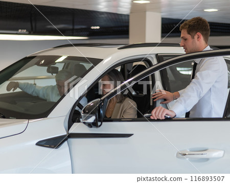 A gray-haired Caucasian woman is sitting behind the wheel of a car. Car dealership salesman consulting mature married couple. A gray-haired Caucasian woman is sitting behind the wheel of a car. Car dealership salesman consulting mature married couple. 116893507
