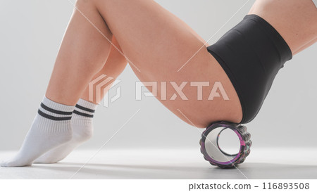 Faceless woman doing roller exercise for myofascial release on white background. Self-massage of thighs. Faceless woman doing roller exercise for myofascial release on white background. Self-massage of thighs. 116893508