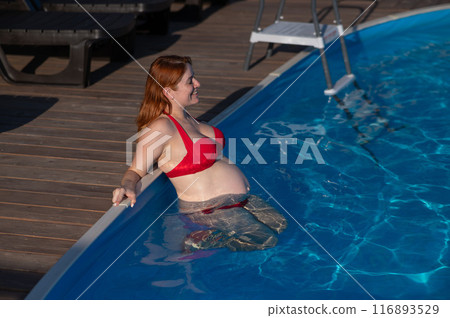 A red-haired pregnant woman in a red bikini is resting in the pool, leaning on the side. A red-haired pregnant woman in a red bikini is resting in the pool, leaning on the side. 116893529