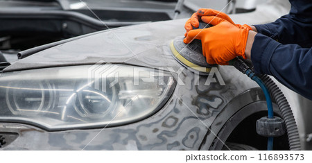 A mechanic sands the putty on a car body with a machine. Repair after an accident. A mechanic sands the putty on a car body with a machine. Repair after an accident. 116893573