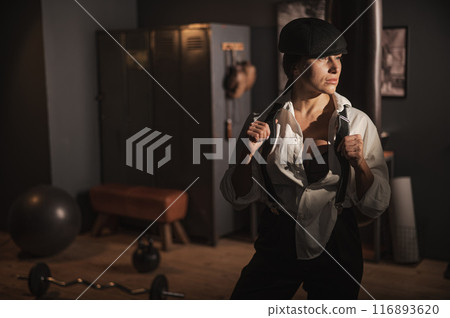 Caucasian woman in a Peaky Blinders style men's suit posing in a retro gym. 116893620