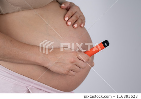 A pregnant woman smokes a vape. A girl holds an electronic cigarette against the background of her bare tummy. Copy space.  116893628