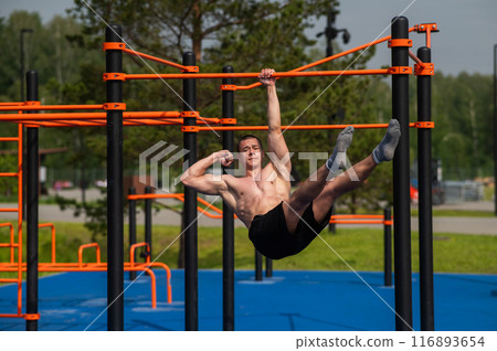 Young muscular man hanging by one hand on a horizontal bar outdoors. Young muscular man hanging by one hand on a horizontal bar outdoors. 116893654