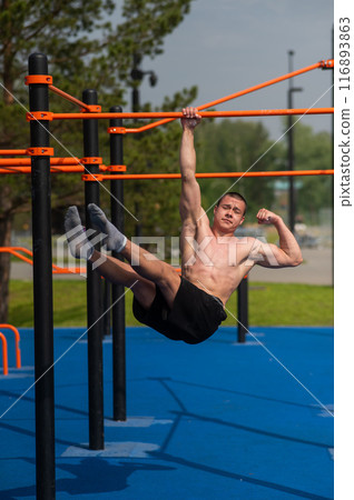 Young muscular man hanging by one hand on a horizontal bar outdoors. Vertical photo.  116893863