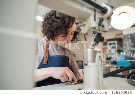 A woman tanner sews a leather belt on a sewing machine.  116893926