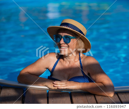 Portrait of an elderly woman in a hat and blue sunglasses swims in the pool. Vacation in retirement.  116893947