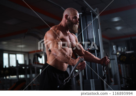 Bald Caucasian bodybuilder training chest using cable crossover in gym.  116893994